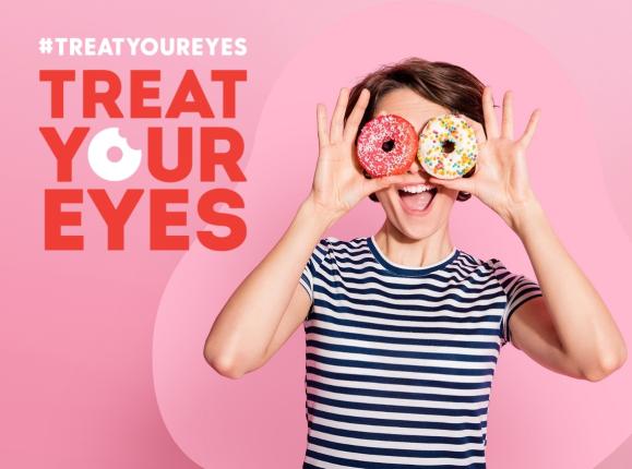 Image of young woman in black and white striped t-shirt, smiling and holding donuts over her eyes, with a pink background and red type stating 'Treat Your Eyes'